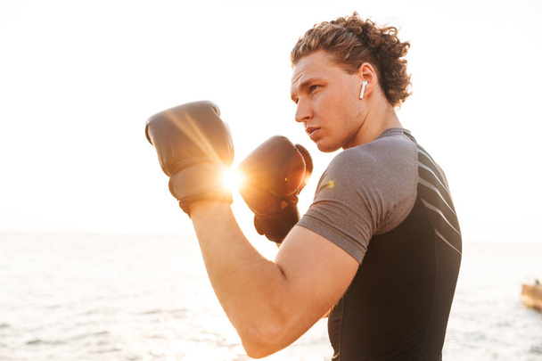 Photo of young handsome sports man boxer outdoors on the beach boxing in gloves. - Фото, изображение