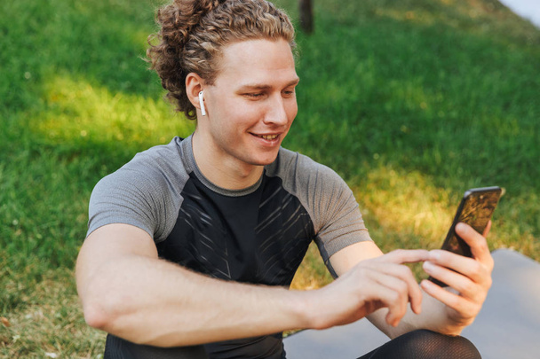 Smiling young sportsman listening to music with earphones while sitting on a fitness mat outdoors, holding mobile phone - Foto, Bild