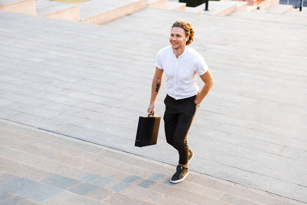 Happy curly business man with briefcase walking on stairs while holding arm in pocket - Foto, immagini