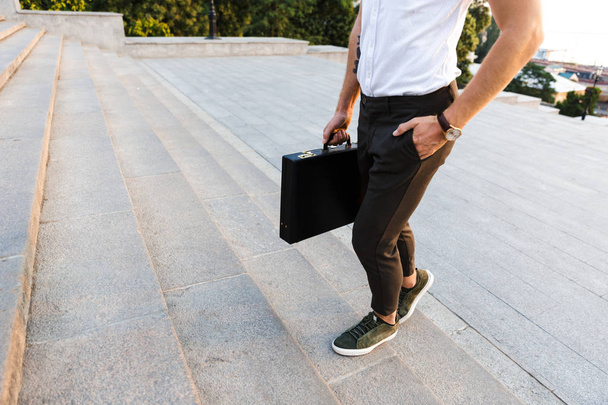Cropped image of young business man with briefcase walking on stairs while holding arm in pocket - Фото, зображення