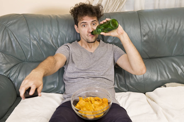 Excited young man with the remote and beer and chips on the couch - Foto, afbeelding