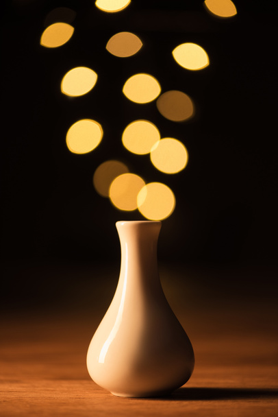 close up view of white vase and yellow bokeh lights on black background - Foto, afbeelding