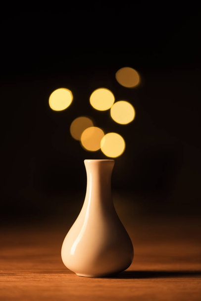 close up view of white vase and yellow bokeh lights on black background - Фото, изображение