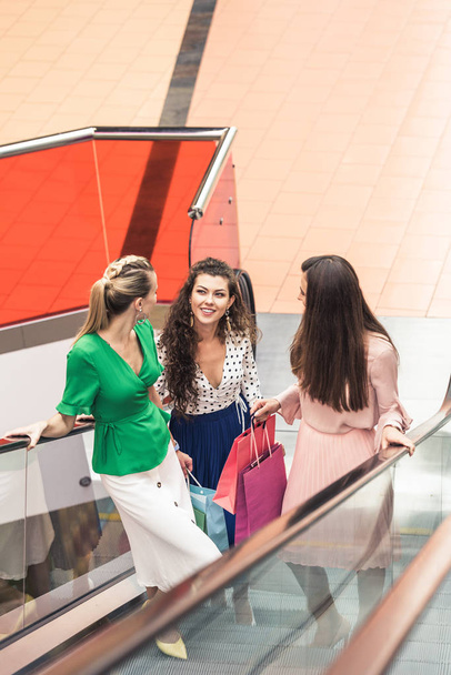 Blick aus der Vogelperspektive auf lächelnde stilvolle Mädchen, die Papiertüten in der Hand halten und auf einer Rolltreppe im Einkaufszentrum sprechen - Foto, Bild