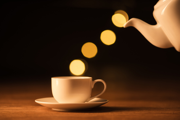 close up view of white teapot, cup of coffee and bokeh lights on black backdrop - Fotó, kép