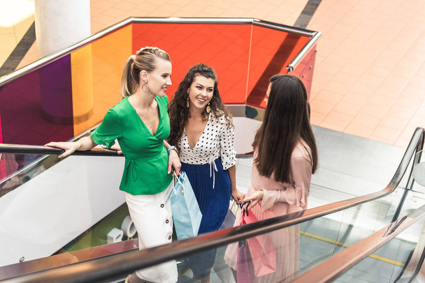 vista de ángulo alto de las mujeres jóvenes sonrientes sosteniendo bolsas de compras y de pie juntos en escaleras mecánicas
 - Foto, Imagen
