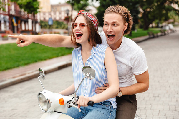 Iloisen nuoren pariskunnan muotokuva ajamassa moottoripyörällä yhdessä kaupungin kadulla, osoittaen sormella
 - Valokuva, kuva
