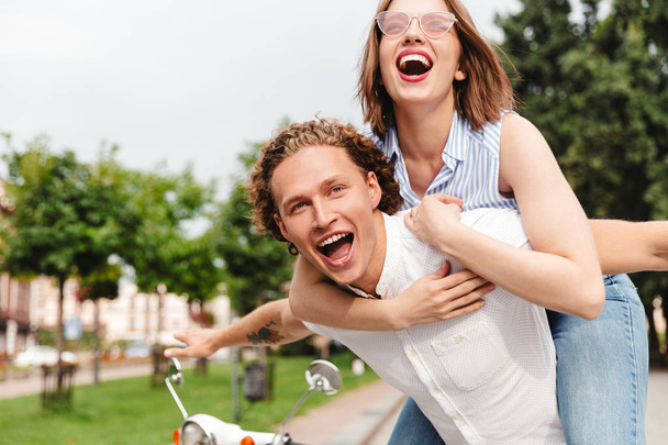 Alegre joven pareja divirtiéndose junto con scooter y mirando a la cámara mientras está en el parque
 - Foto, imagen