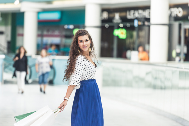красивая стильная молодая женщина держит сумки с покупками и улыбается перед камерой в торговом центре
 - Фото, изображение