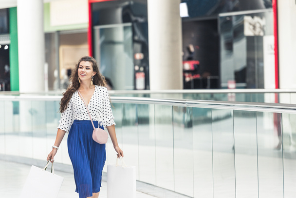 beautiful smiling young woman holding paper bags and looking away while walking in shopping mall - Фото, изображение
