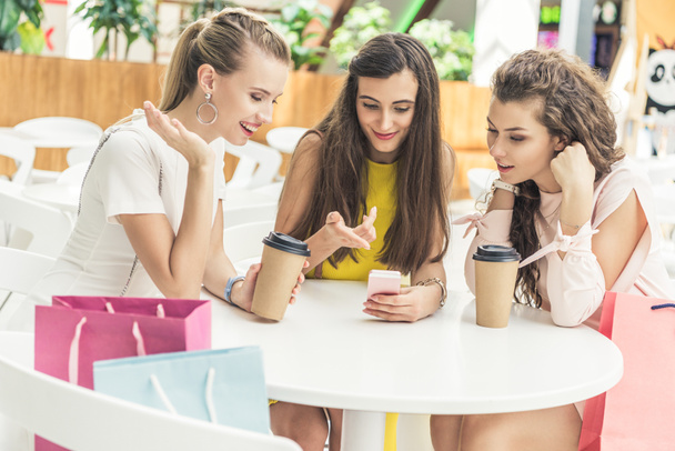 mujeres jóvenes sonrientes usando teléfono inteligente y beber café de tazas de papel en la cafetería en el centro comercial
 - Foto, imagen