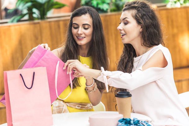 smiling young women looking into shopping bag while sitting at table in shopping mall - Foto, Bild