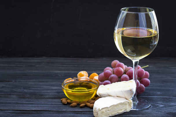 A glass of white wine with cheese cuts, figs, nuts, honey, grapes on a dark rustic wooden boards background. Top view - Valokuva, kuva