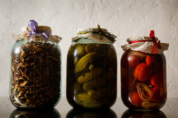 trois grands bocaux en verre avec des tomates en conserve maison, des concombres et des pommes séchées se tiennent en rangée sur la table de cuisine contre le mur gris
 - Photo, image