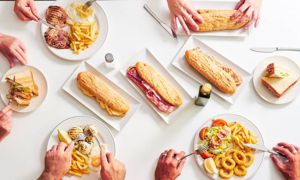 Overhead view of friends sitting at a restaurant table. Grilled steaks, French fries, salad, sandwiches and fried squid on white table. - Photo, Image