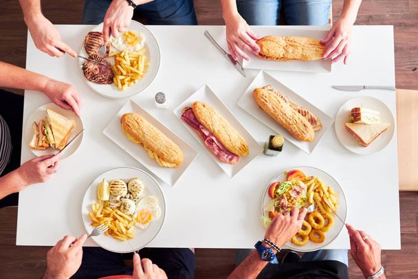 Overhead view of friends sitting at a restaurant table. Grilled steaks, French fries, salad, sandwiches and fried squid on white table. - Fotoğraf, Görsel