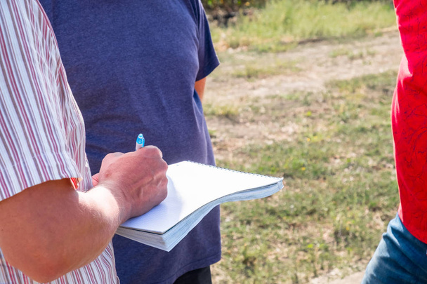 a man in a colored shirt with a notebook in his hands writes with a pen, discusses with another person in the field in the summer agronomist - Foto, Imagen