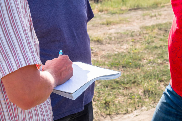 a man in a colored shirt with a notebook in his hands writes with a pen, discusses with another person in the field in the summer agronomist - Φωτογραφία, εικόνα
