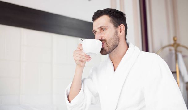 Handsome brunette bearded man stands before bright window and drinks coffee - Foto, imagen