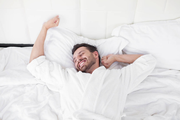 Cheerful young man is waking up after sleeping in the morning. He is yawing and stretching his arms up. His eyes are closed with relaxation. He is lying in the bed - Foto, imagen
