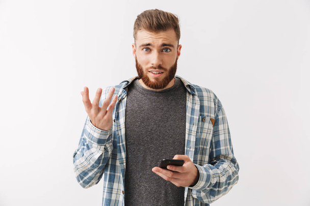 Portrait of a frustrated young bearded man standing isolated over white, holding mobile phone - Fotó, kép