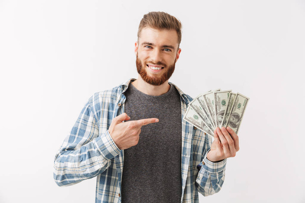 Portrait of a cheerful young bearded man standing isolated over white, showing money banknotes, pointing - Foto, Bild
