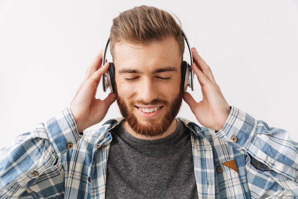 Pleased bearded man in shirt and headphones listening music with closed eyes over grey background - Photo, image