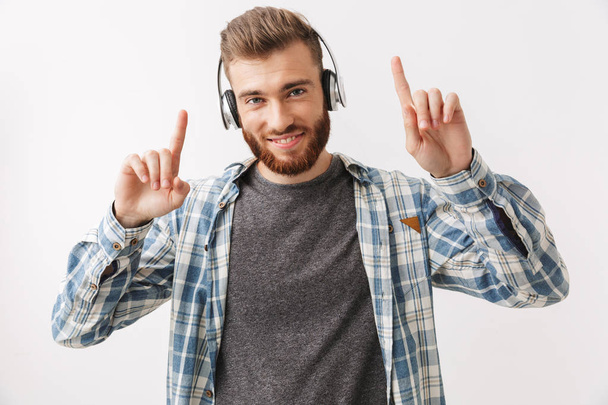Happy bearded man in shirt and headphones listening music and showing his forefingers while looking at the camera over rey background - Photo, image