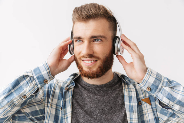 Pleased bearded man in shirt and headphones listening music while looking away over grey background - 写真・画像