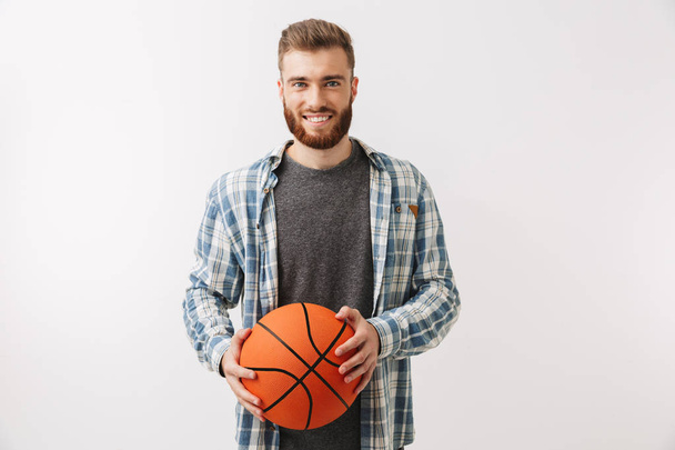 Smiling bearded man in shirt holding basketball and looking at the camera over grey background - Foto, afbeelding