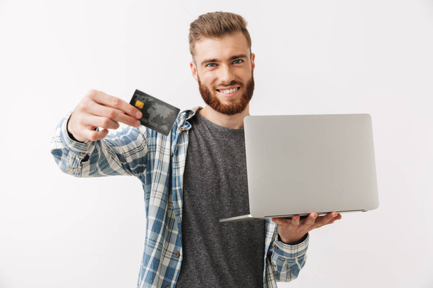 Portrait of a confident young bearded man standing isolated over white, holding laptop and showing credit card - 写真・画像