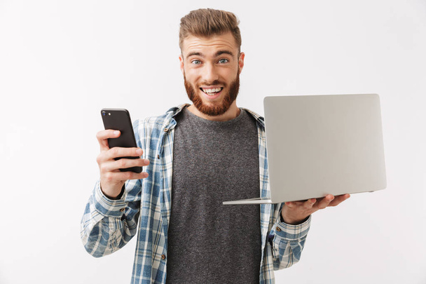 Portrait of a confident young bearded man standing isolated over white, holding laptop and mobile phone - Фото, изображение