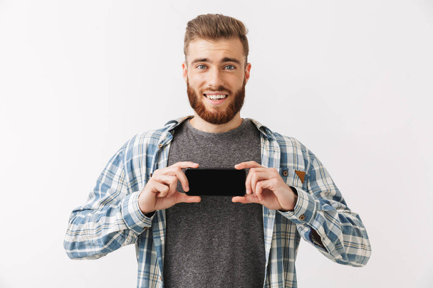 Portrait of a confident young bearded man standing isolated over white, showing blank screen mobile phone - Фото, зображення