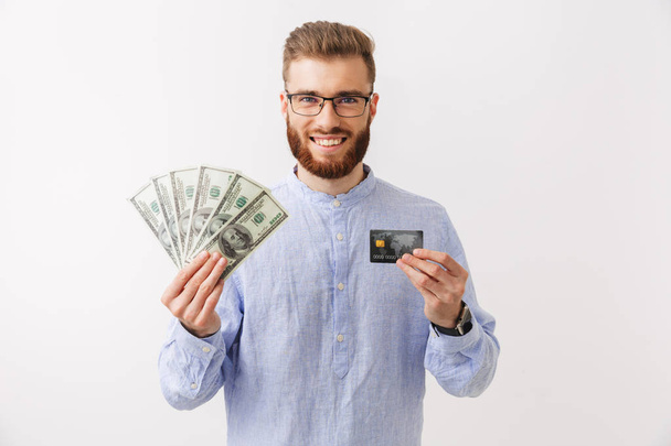 Portrait of a smiling young bearded man standing isolated over white, showing money banknotes and credit card - Fotoğraf, Görsel