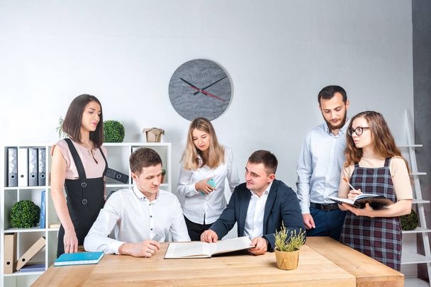 Theme is business and teamwork. A group of young Caucasian people office workers holding a meeting, briefing, working with papers and documents in a light office office around a wooden table. - Photo, Image