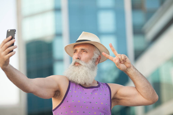 Old active bearded man in hat taking selfie with mobile phone on street. - Photo, image