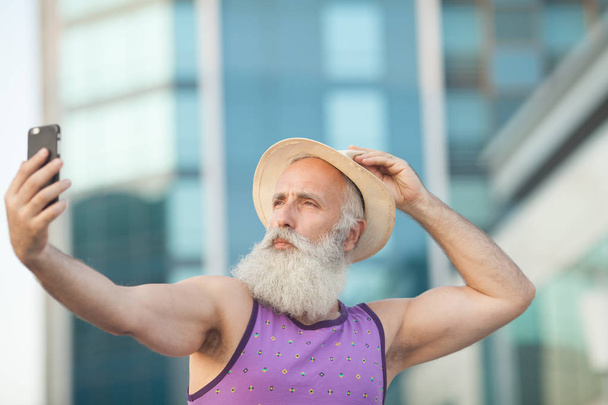Старый бородатый мужчина в шляпе делает селфи с мобильным телефоном на улице
. - Фото, изображение