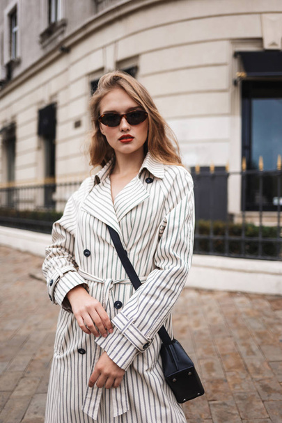Young attractive woman in striped trench coat with little black cross bag thoughtfully looking in camera walking around cozy city street - Foto, Bild