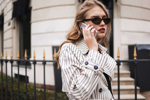 Молода серйозна жінка в смугастій траншеї пальто задумливо говорить по мобільному телефону, ходячи навколо затишної міської вулиці
 - Фото, зображення