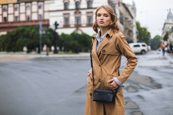 Mujer atractiva joven en abrigo de trinchera con pequeño bolso cruzado negro mirando soñadoramente en cámara pasar tiempo en la acogedora calle de la ciudad
 - Foto, imagen