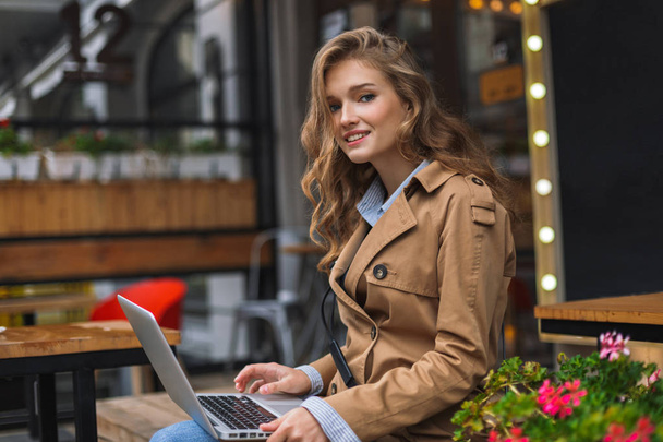 schönes lächelndes Mädchen im Trenchcoat, das verträumt in die Kamera blickt und mit Laptop auf Knien Zeit im Freien auf der Caféterrasse verbringt - Foto, Bild