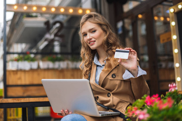 Mujer bastante sonriente en gabardina mirando soñadoramente en la cámara con tarjeta de crédito en la mano sentado con el ordenador portátil en las rodillas al aire libre en la terraza de la cafetería
 - Foto, Imagen