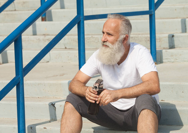 closeup grey bearded old man in white vest ready to do morning exercises drink water bottle - Fotografie, Obrázek