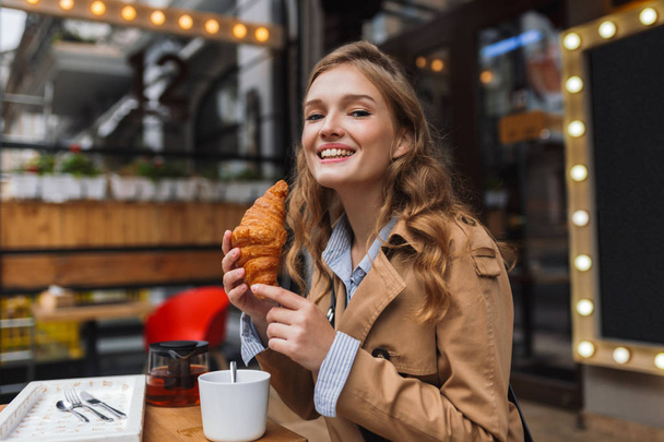 Young smiling woman in trench coat happily looking in camera holding croissant in hand spending time outdoor at cozy cafe terrace - Photo, Image