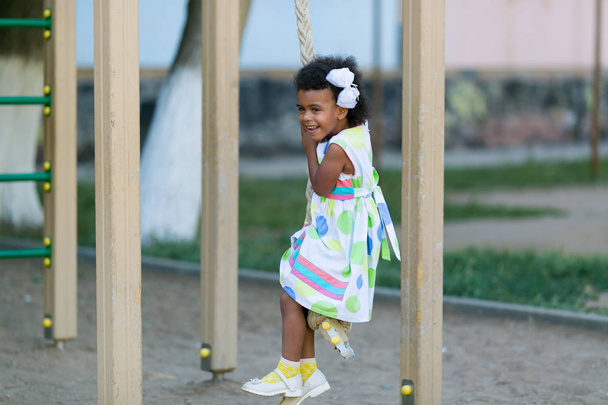 African American girl plays on a sports playground. The girl is hanging on the sports rings. - Fotó, kép