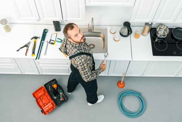 vysoký úhel pohled pohledný instalatér drží nastavitelný klíč a při pohledu na fotoaparát v kuchyni - Fotografie, Obrázek