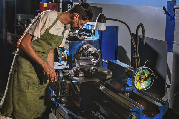 vue latérale du travailleur de fabrication masculin dans le tablier et les lunettes de protection à l'aide de la machine-outil à l'usine
 - Photo, image