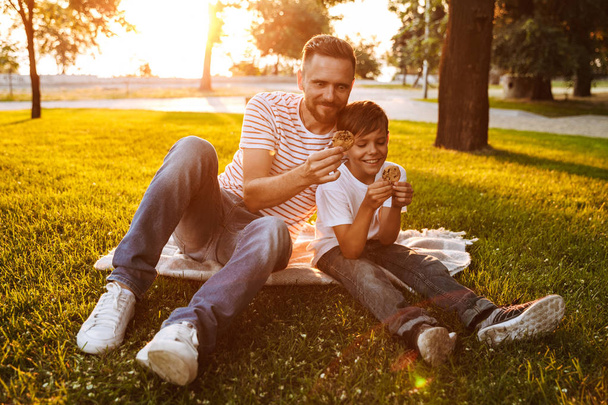 Cheerful father spending time with his little son at the park, eating cookies - Fotoğraf, Görsel