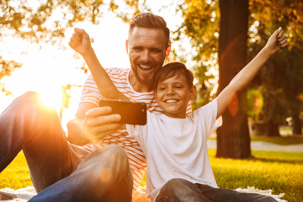 Image of happy young man father have a rest with his son outdoors in park play games with mobile phone. - Foto, afbeelding