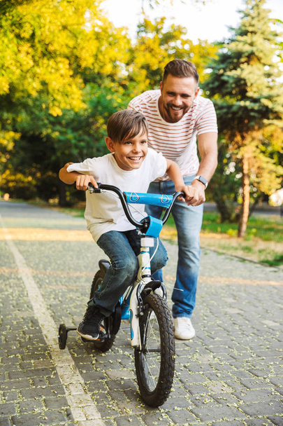 Smiling father and his son having fun together at the green park, teaching son how to ride a bicycle - Фото, изображение